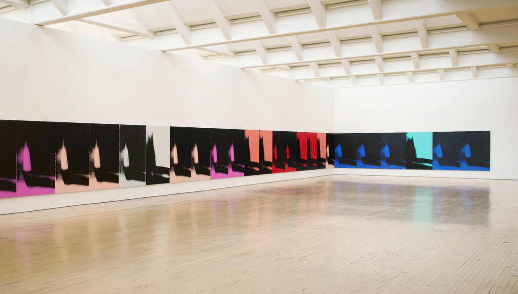 Twenty-three canvases hang edge to edge and low to the floor along a wall and in the corner of a gallery.  Each canvas features an identical abstract mark painted in black, gray, red, green, yellow, peach, or blue.